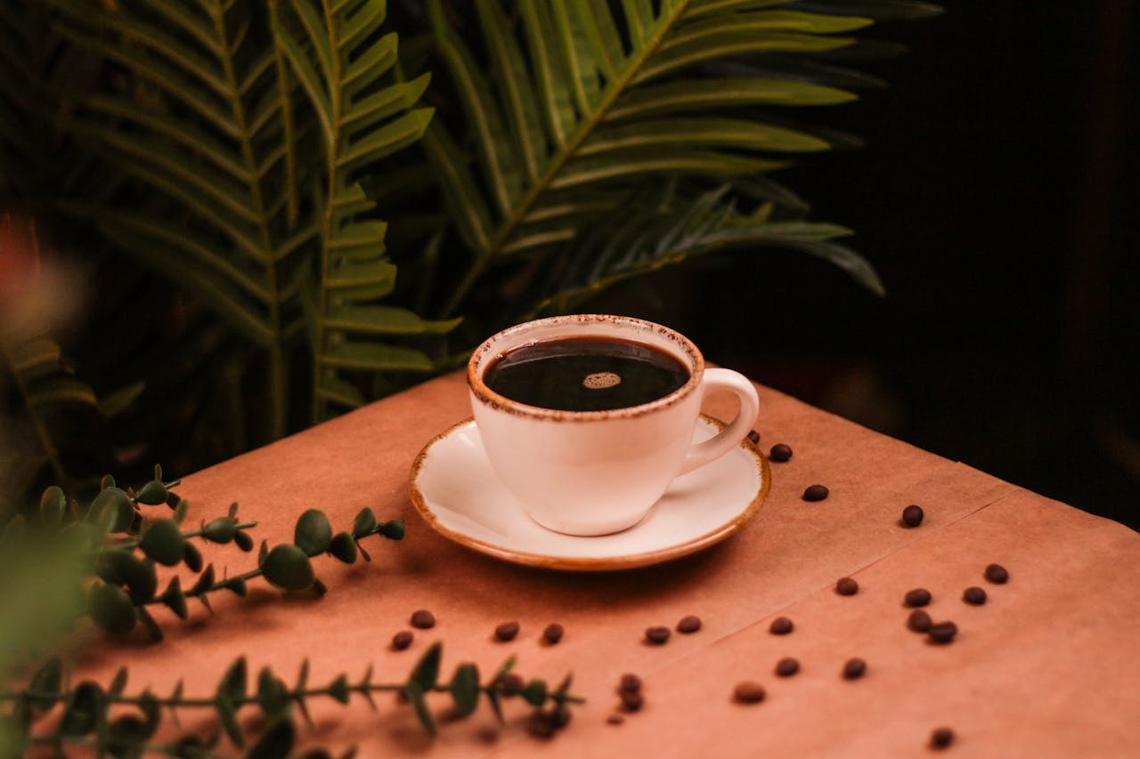 gallery-6 A warm coffee cup on a wooden table with plants and coffee beans, creating a cozy atmosphere.
