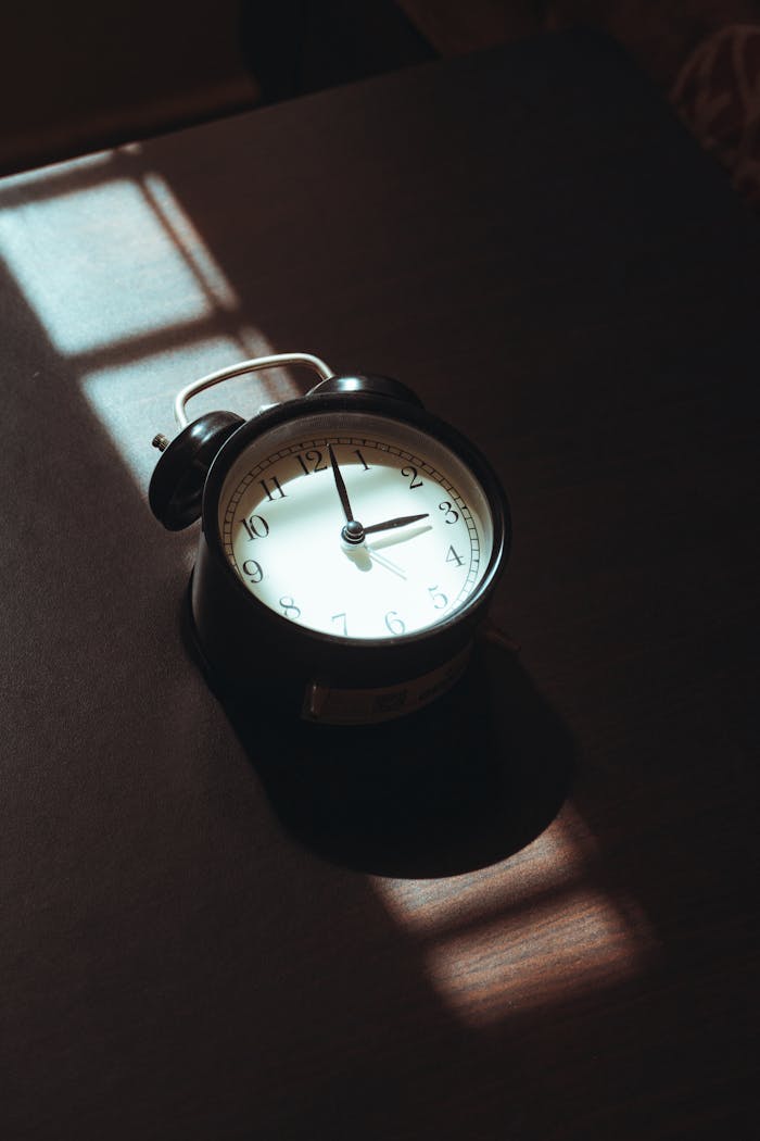 gallery-1 A vintage black analog clock on a wooden surface bathed in soft morning light, creating an artistic shadow play.