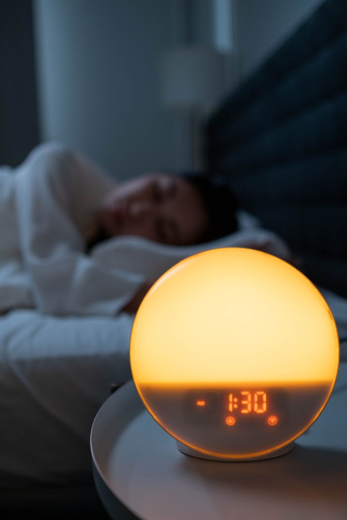 gallery-2 Warm orange glow of alarm clock illuminating a sleeping person in dimly lit bedroom.